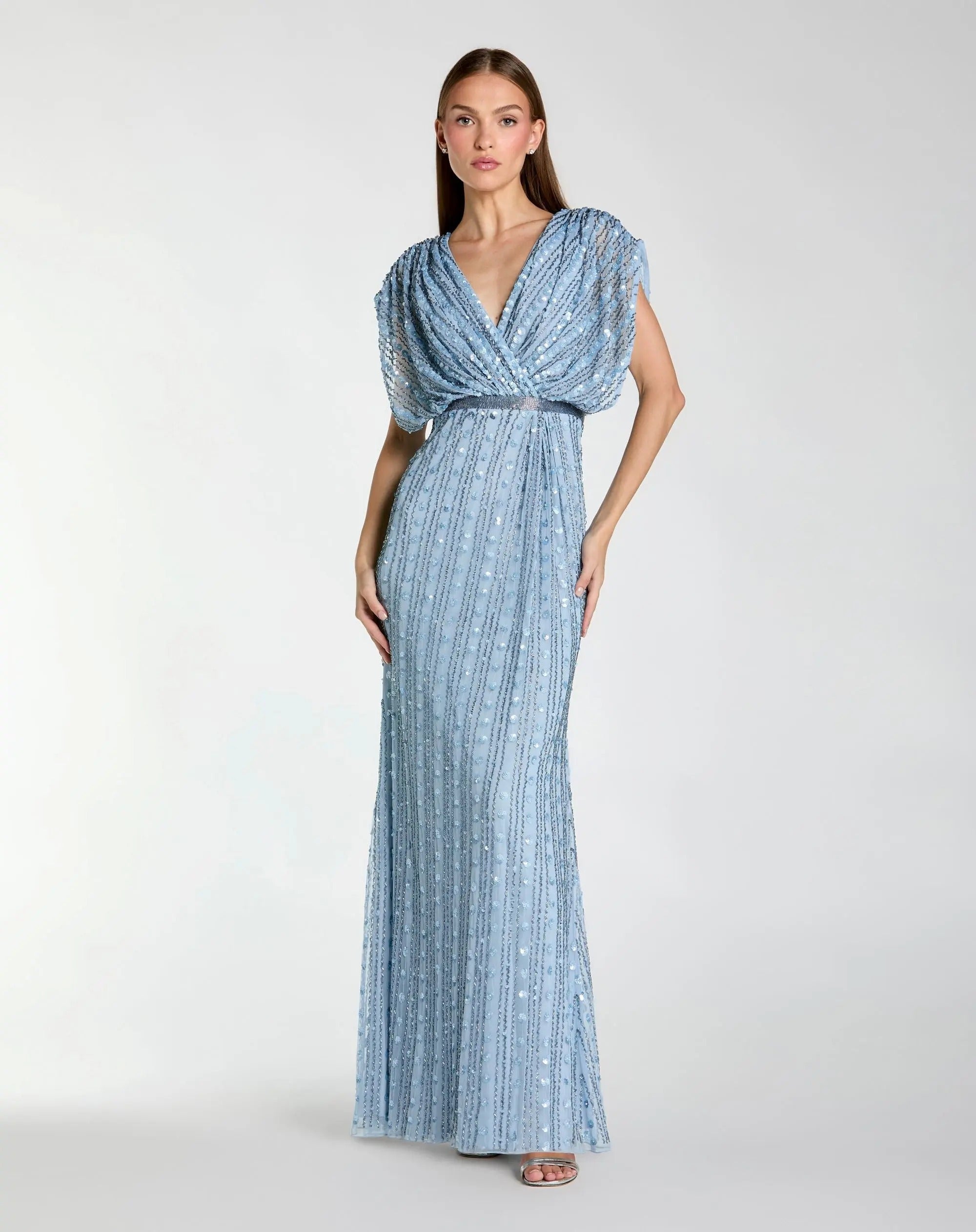 A woman stands against a plain light background wearing the Mac Duggal 23081 Long Formal Dress in periwinkle—a floor-length gown adorned with sequin embellishments, featuring short sleeves and a deep V-neckline.